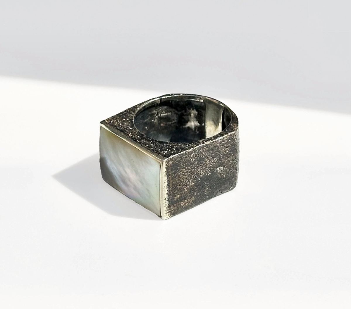 Anillo de plata texturizado con nácar, bajo luz de sol dura sobre blanco.
