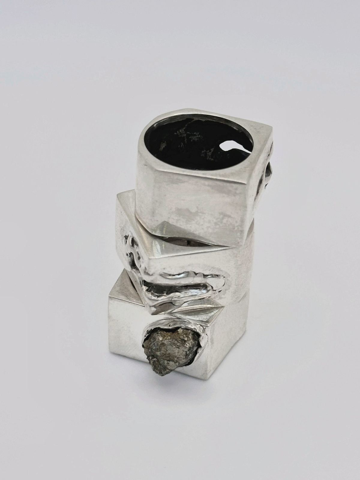 Anillos cuadrados de plata apilados, uno con pirita en bruto, sobre gris claro.