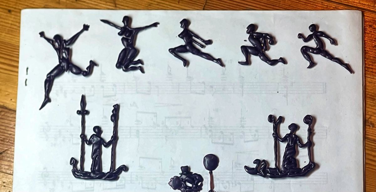 Siluetas de bailarines y guerreros talladas en cera oscura sobre una partitura.